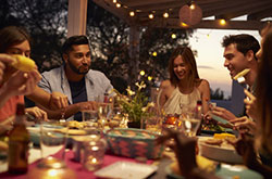 Freunde sitzen abends auf der Terasse und essen ein Festmahl, während Lichterketten das Geschehen beleuchten.